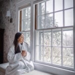 A woman in a plush robe sits along a window bench looking out the window at the landscape.