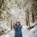 A woman stands in a winter forest covered in snow, with trees all around, throwing snow into the air