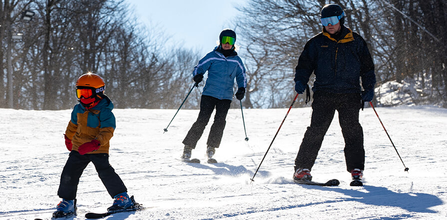 A family bundled up in skiing gear skiis down a small snowy slope together