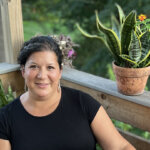 Julie sits looking at the camera on a bright, summer day with plants surrounding her