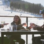 A family of two adults and one child sit around a table eating lunch at the base of the snowy ski slopes of Bousquet Moutain.