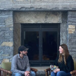 A man and a woman look at each other talking, sitting in front of the large stone fireplace at the Greylock Glen