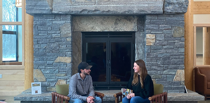 A man and a woman look at each other talking, sitting in front of the large stone fireplace at the Greylock Glen