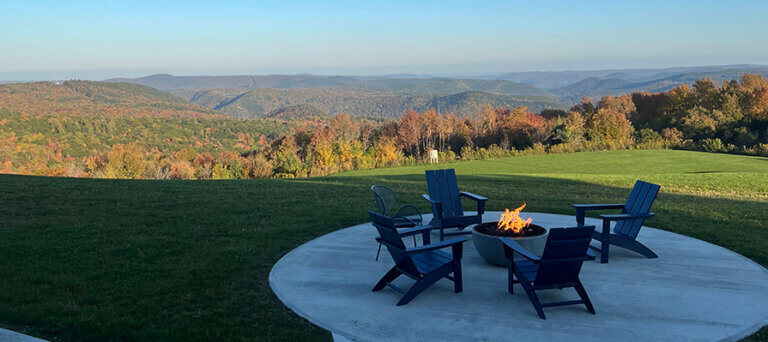 Blue Vista Motor Lodge_credit Cathleen King (2) 895×400 Four blue lounge chairs circle around a fire pit outside the Blue Vista Motor Lodge. In the background the trees and mountains filled with colorful foliage.