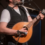 A man sings into a microphone while strumming a guitar