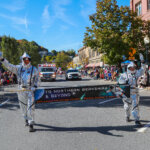 Two adults march in a parade wearing astronaut costumes, carrying a banner