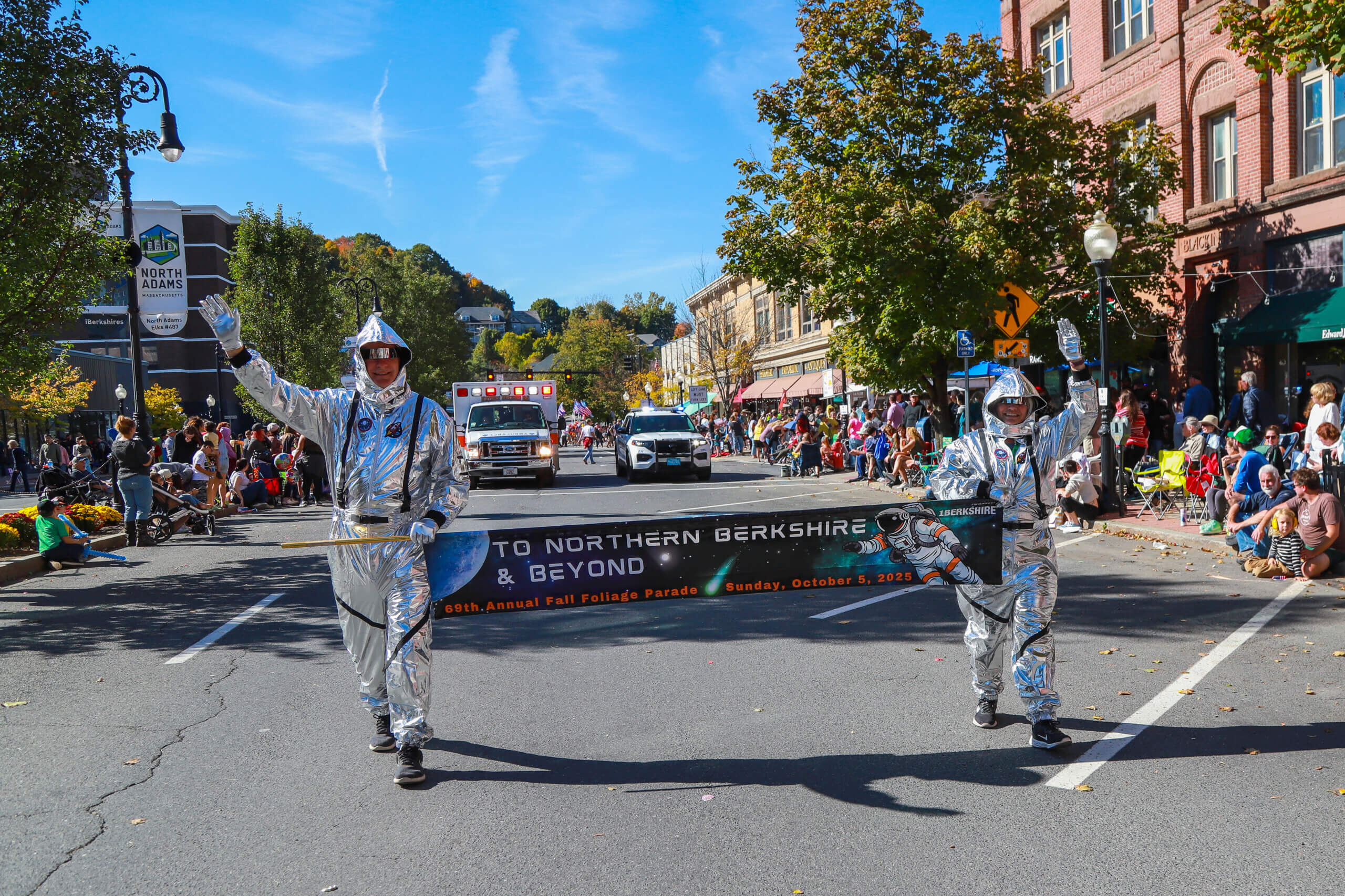 Two adults march in a parade wearing astronaut costumes, carrying a banner