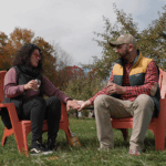 p58 2022 FALL_Great Sky Media_4 – Hilltop (1) 895×400 A couple holds hands as they sip cider in a fall apple orchard