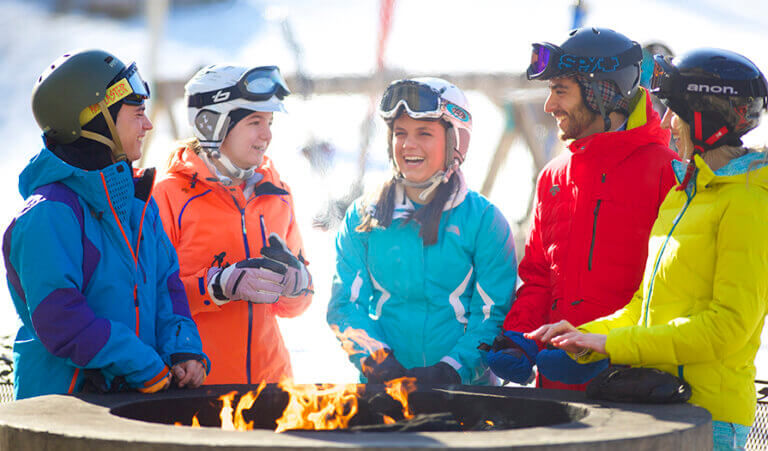 A group of skiiers stands around a fire pit at Jiminy Peak laughing.