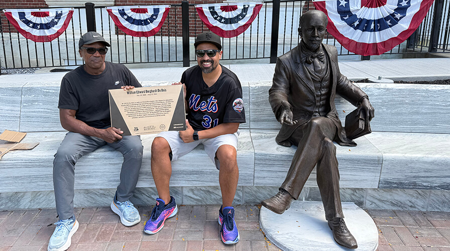 Two men sit next to the newly unveiled WEB DuBois statue holding a plaque
